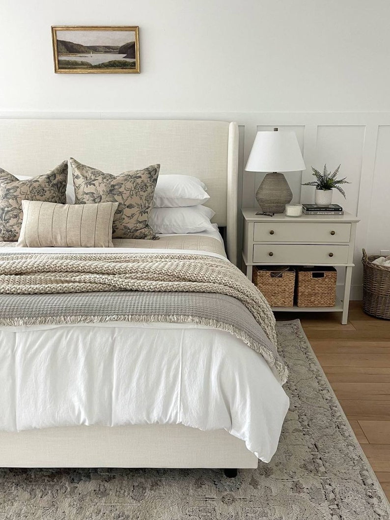May include: A white bed with a beige headboard and a white duvet cover. The bed has a beige knitted throw blanket and a gray throw blanket with fringe. There are two pillows with a floral pattern and a beige pillow with stripes on top of the bed. A white lamp with a beige shade is on a nightstand next to the bed. The nightstand has two drawers and two wicker baskets. A small potted plant is on the nightstand.