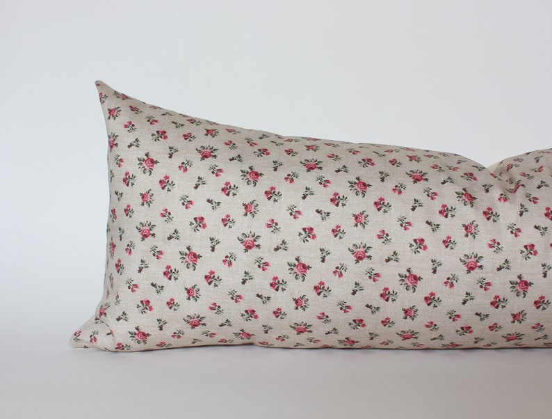 May include: A long, rectangular pillow with a beige linen fabric cover featuring a small-scale floral pattern of pink roses and green leaves.