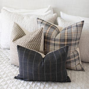 May include: A selection of decorative cushions in a range of patterns and colours. The cushions include a grey and white plaid design, a grey and white striped cushion, and a patterned cushion in a neutral colour scheme. The cushions are arranged on a white quilted bedspread.