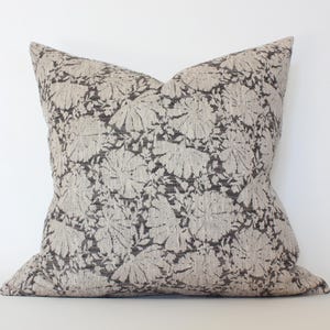 May include: Square cushion with a neutral colour scheme. It features a repeating pattern of stylised floral shapes in beige and dark grey. This soft furnishing item is suitable for a living room or bedroom. The cushion is a decorative item.