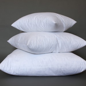 Puede incluir: Tres almohadas blancas apiladas una encima de la otra sobre un fondo gris.