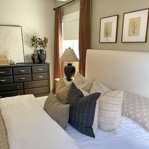 K&ouml;nnte beinhalten: Ein wei&szlig;es Bett mit einem wei&szlig;en Kopfteil und einem wei&szlig;en Bettbezug. Auf dem Bett liegen mehrere Kissen, darunter ein schwarz-wei&szlig; gestreiftes Kissen, ein beiges Kissen und ein braunes Kissen. Neben dem Bett steht eine Lampe auf dem Nachttisch.