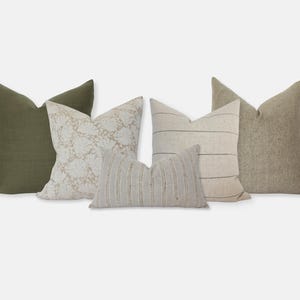 Op de afbeelding: Vijf decoratieve sierkussens in verschillende texturen en patronen. De kussens omvatten effen olijfgroen, crème met een bloemmotief, strepen en getextureerd beige. De kussens zijn gerangschikt op een lichtgrijze achtergrond.