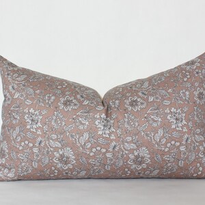 May include: A blush pink lumbar pillow with a white floral pattern. The pillow is made of a soft, woven fabric.