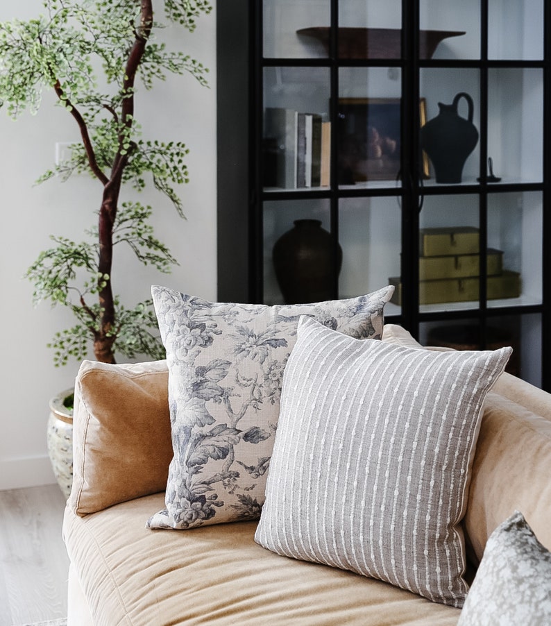 May include: A tan couch with three throw pillows. The pillows are patterned with floral designs and stripes. The couch is in front of a black metal shelving unit with glass shelves.