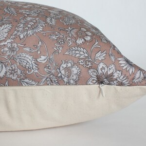 May include: A blush pink pillow with a floral pattern in shades of gray and white. The pillow has a zipper closure on the bottom.