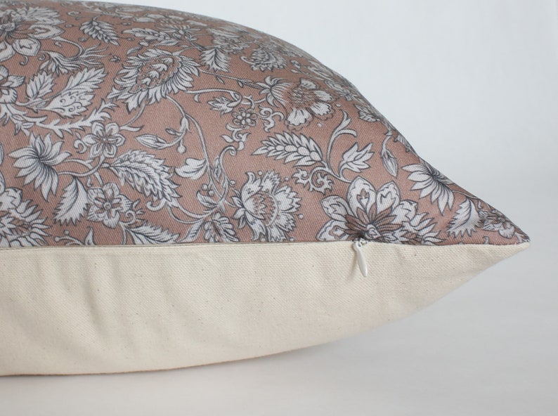 May include: A blush pink pillow with a floral pattern in shades of gray and white. The pillow has a zipper closure on the bottom.