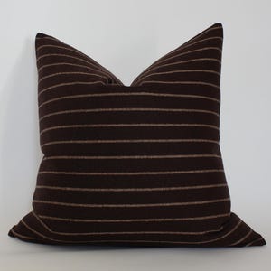 May include: A square decorative pillow with a dark brown background and horizontal tan stripes. The pillow is made of a soft fabric and is designed for home decor. The pillow is approximately 18 inches square.