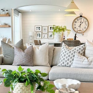 May include: A living room with a gray couch and a variety of throw pillows. The couch is in front of a large window and a white wall with framed pictures. There is a coffee table in front of the couch with a potted plant, a white bowl, and a stack of books. The books have the word "ABODE" on the cover.