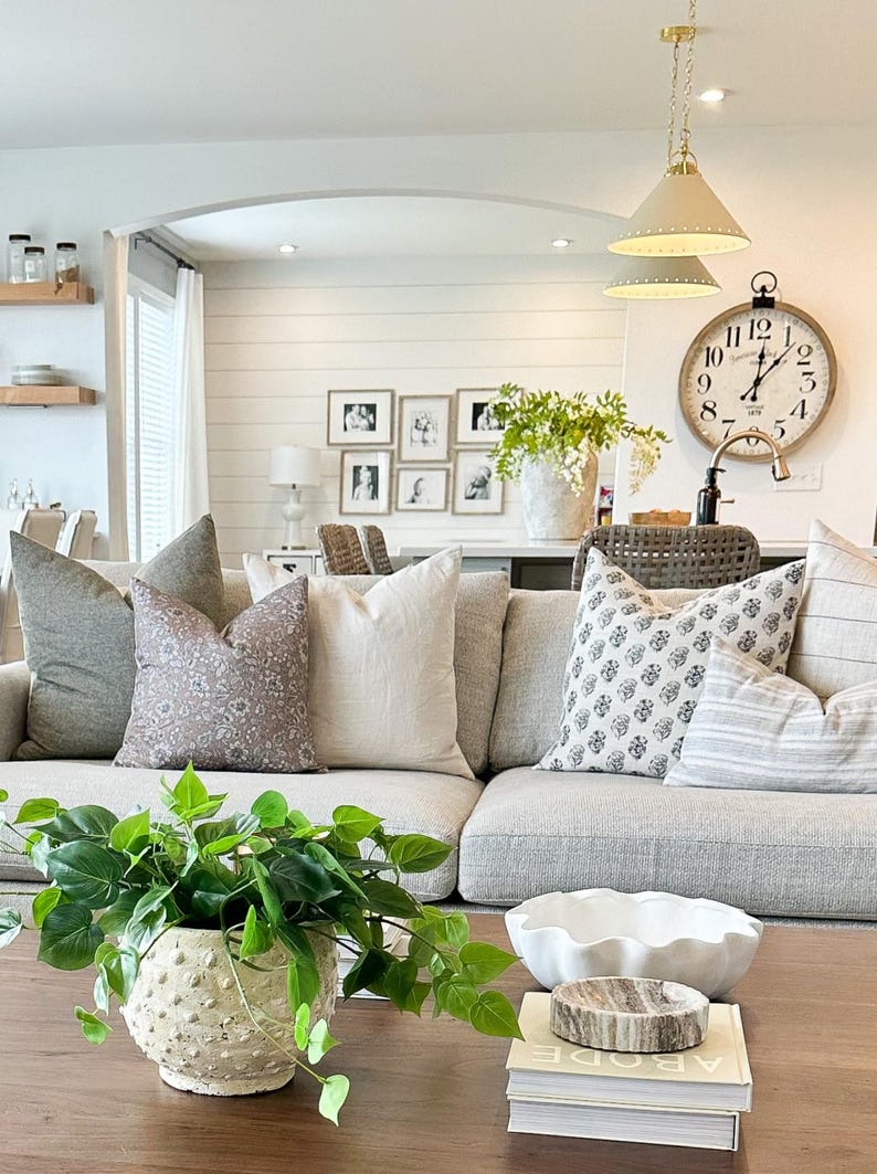 May include: A living room with a gray couch and a variety of throw pillows. The couch is in front of a large window and a white wall with framed pictures. There is a coffee table in front of the couch with a potted plant, a white bowl, and a stack of books. The books have the word "ABODE" on the cover.