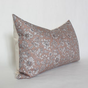 May include: A rectangular decorative pillow with a floral pattern. The pillow features a dusty rose-colored fabric with a white and gray floral design on the front. The back and sides of the pillow are a solid cream color.