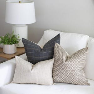May include: Four decorative throw pillows in neutral tones rest on a white sofa. The pillows feature various textures and patterns, including a plaid, a geometric design, and a woven texture. A white lamp and potted plant are in the background.