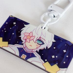Puede incluir: Un llavero rectangular con un personaje de estilo anime con cabello blanco y una flor rosa. El fondo es azul oscuro con estrellas blancas. El llavero tiene un cierre blanco.