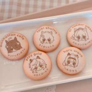 May include: Six round cookies with cartoon character illustrations and Japanese text. The cookies are light brown with brown illustrations and text. The cookies are arranged on a white rectangular plate.