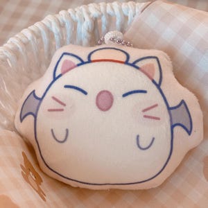 May include: A plush, white, cartoon-style character keychain with blue and purple accents. The character has a smiling face, small wings, and a red hat. The keychain is resting in a white basket with a pink and white checkered cloth.