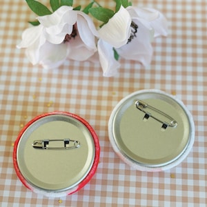 May include: Two round pinback buttons with safety pin closures. One button has a red and white patterned edge, the other has a white and pink patterned edge. The buttons are on a tan and white checkered surface with white flowers.