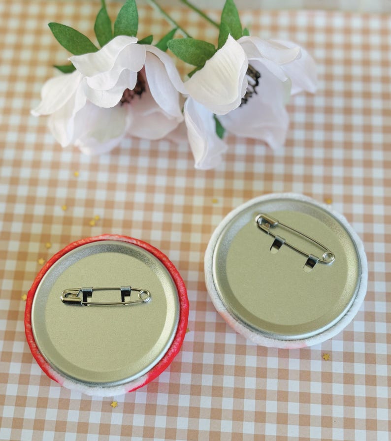 May include: Two round pinback buttons with safety pin closures. One button has a red and white patterned edge, the other has a white and pink patterned edge. The buttons are on a tan and white checkered surface with white flowers.