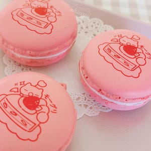 May include: Three pink macarons with a white filling and a red cartoon design on top. The macarons are arranged on a white doily and a light pink tray. The design appears to be a cat with a strawberry.