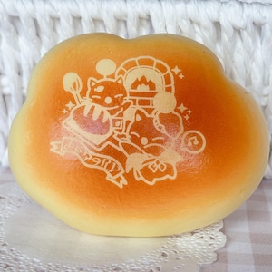 May include: A golden-brown, cloud-shaped pastry with a detailed white illustration of cartoon cats, a bread oven, and text. The pastry sits on a white doily and a checkered surface, with a white woven basket in the background.