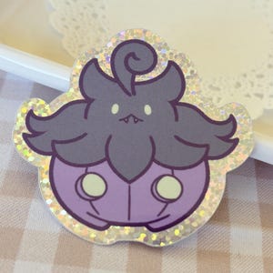 May include: A holographic sticker featuring a cartoon character with a dark gray head and wings, a purple body, and two yellow circles. The sticker has a glittery border and is set against a checkered background.