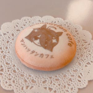 May include: A round, light-colored cookie with a brown image of a cat and the words "Manhattan cafe" in Japanese. The cookie sits on a white, lacy paper doily.