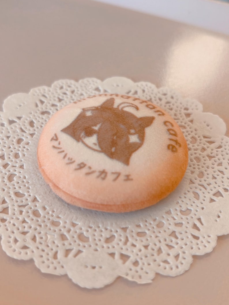 May include: A round, light-colored cookie with a brown image of a cat and the words "Manhattan cafe" in Japanese. The cookie sits on a white, lacy paper doily.