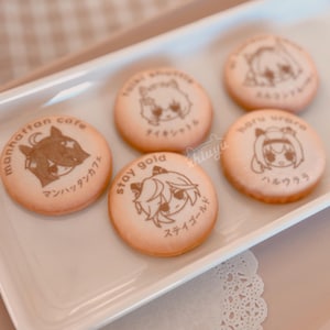 May include: A white rectangular tray holds six round cookies. Each cookie has a light brown color and features a different cartoon character design with Japanese text. The cookies are arranged on a white tray with a decorative lace doily.