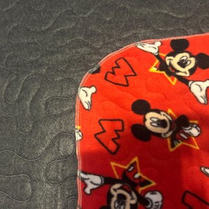 May include: A red fleece blanket featuring a pattern of Mickey Mouse characters and yellow stars. The blanket has a gray, quilted backing. The edge is finished with a black stitch.