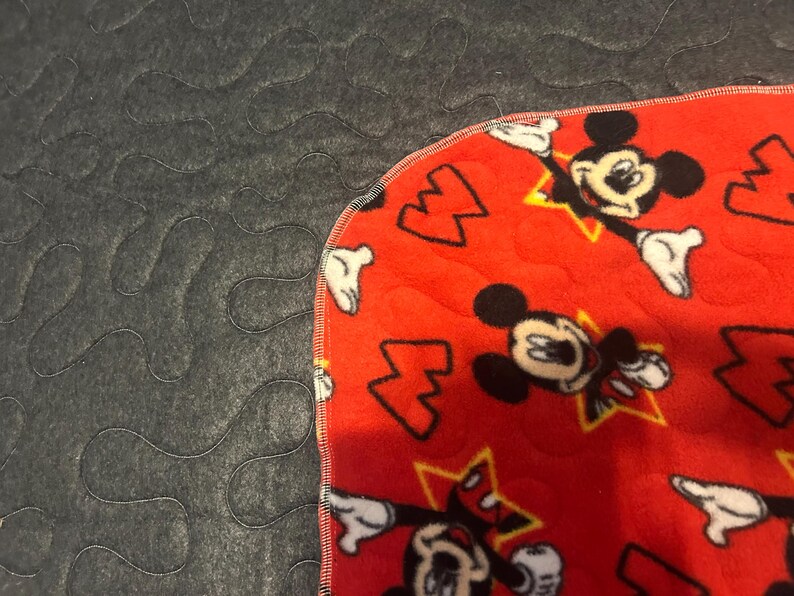 May include: A red fleece blanket featuring a pattern of Mickey Mouse characters and yellow stars. The blanket has a gray, quilted backing. The edge is finished with a black stitch.
