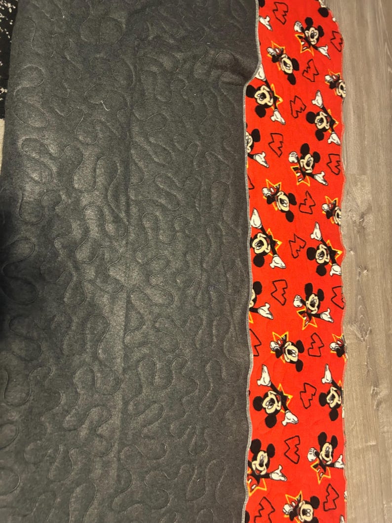 May include: A section of a quilt with a gray, patterned fabric on the left and a red fabric with a Mickey Mouse print on the right. The Mickey Mouse fabric has a white border.