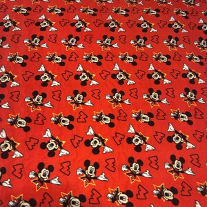 May include: Red blanket featuring a repeating pattern of Mickey Mouse in various poses. Mickey is depicted in black, white, and yellow against a red background. The blanket is square-shaped and appears to be made of a soft material.
