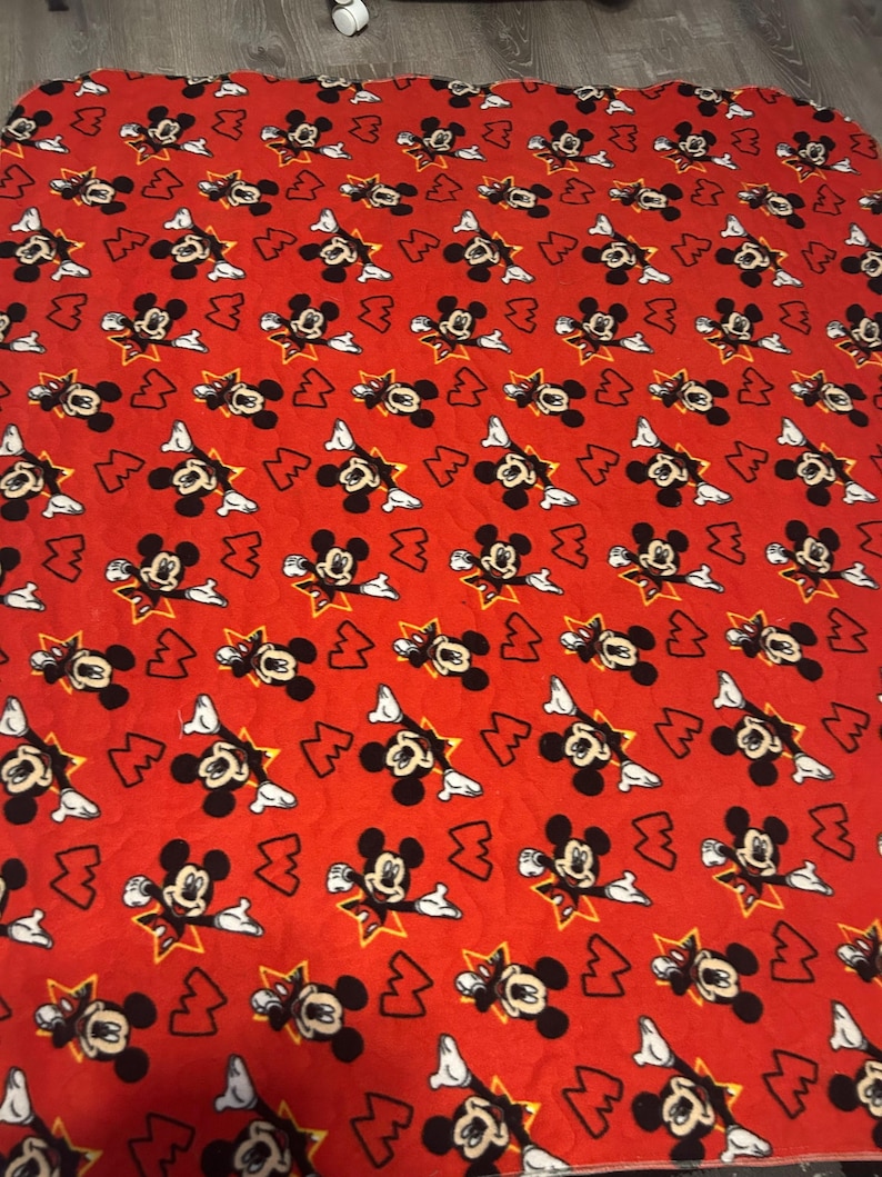 May include: Red blanket featuring a repeating pattern of Mickey Mouse in various poses. Mickey is depicted in black, white, and yellow against a red background. The blanket is square-shaped and appears to be made of a soft material.