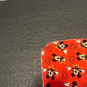 May include: A red fabric with a pattern of Mickey Mouse and lightning bolts. The fabric is on a dark gray textured surface. The Mickey Mouse figures are black and white with yellow stars.
