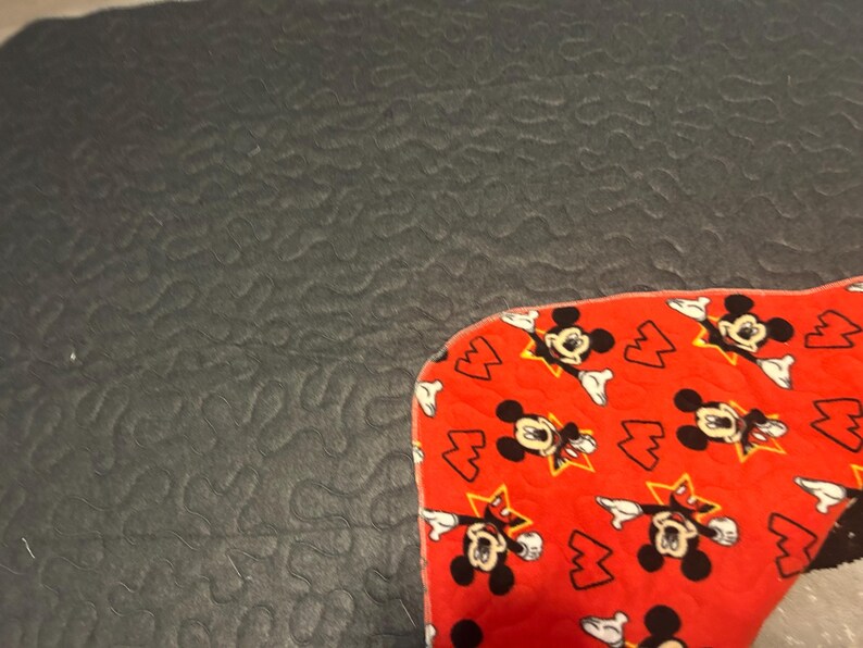 May include: A red fabric with a pattern of Mickey Mouse and lightning bolts. The fabric is on a dark gray textured surface. The Mickey Mouse figures are black and white with yellow stars.