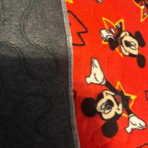 May include: A red fleece blanket featuring a pattern of Mickey Mouse and yellow stars. The blanket is bordered by a dark gray fabric with a quilted design. The two fabrics are sewn together with a gray stitch.