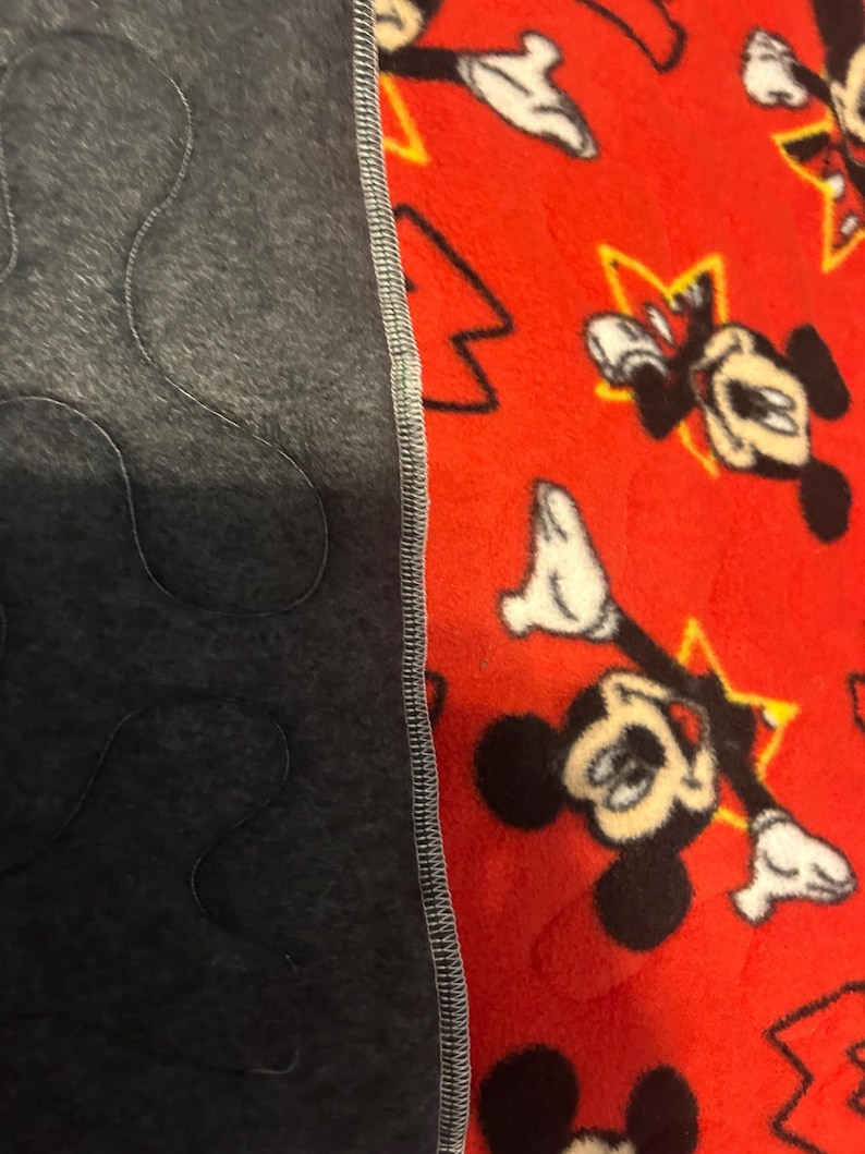 May include: A red fleece blanket featuring a pattern of Mickey Mouse and yellow stars. The blanket is bordered by a dark gray fabric with a quilted design. The two fabrics are sewn together with a gray stitch.