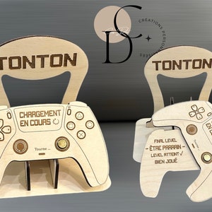 Può includere: Taglio in legno di un controller per videogiochi con il testo "TONTON" e "CHARGEMENT EN COURS" su di esso. Il controller è su un supporto con una maniglia nella parte superiore.