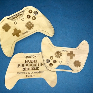 Peut inclure: Deux manettes de jeu en bois avec le texte "TONTON" et "NIVEAU PARRAIN DÉBLOQUÉ" dessus. Les manettes sont en forme de manette de jeu vidéo et ont des boutons et un joystick.
