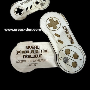 Könnte beinhalten: Ein Holzausschnitt eines Super Nintendo-Controllers mit dem Text "Super Tonton" und "Niveau Parrain Débloqué" darauf. Der Text lautet auch "Acceptes-tu la nouvelle partie?" und enthält die Website www.creas-den.com.