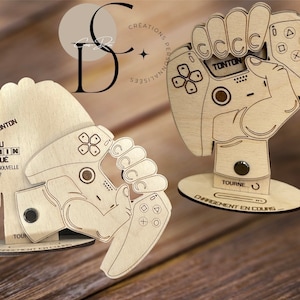 Può includere: Due figurine di legno di mani che tengono un controller per videogiochi. Le figurine sono su una base di legno con il testo "TONTON" e "NIVERU PARRAIN DEBLOQUÉ" sulla base. Il testo "ACCEPTES-TU TA NOUVELLE MISSION?" è anche sulla base.