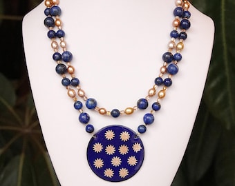 Puede incluir: Collar de dos hilos con cuentas de lapislázuli azul y perlas doradas. El collar presenta un colgante grande, azul oscuro con un patrón de estrellas doradas. El collar se exhibe sobre un busto blanco.
