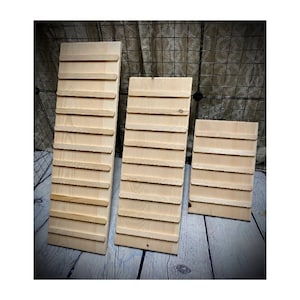 May include: Three unfinished wooden display boards of varying sizes. Each board has horizontal wooden slats attached, creating a tiered design. The boards are made of light-colored wood and are arranged against a neutral background.