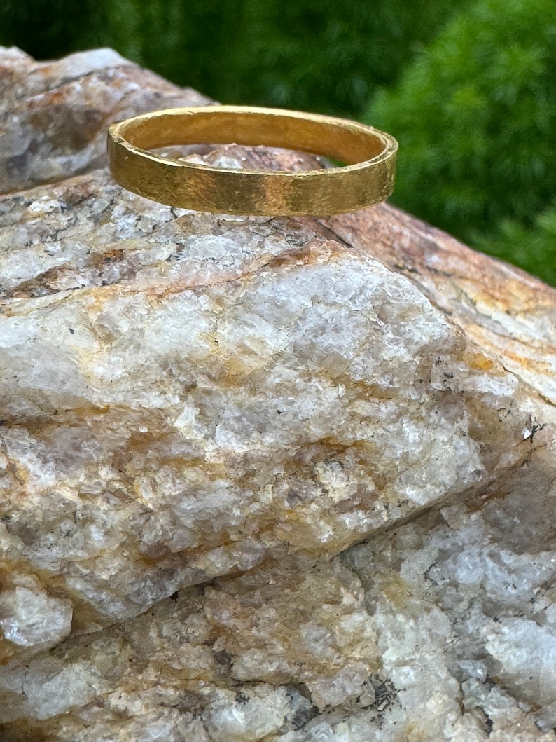 Pode incluir: Um anel de banda de ouro simples com uma textura martelada. O anel est&aacute; sentado em uma rocha.