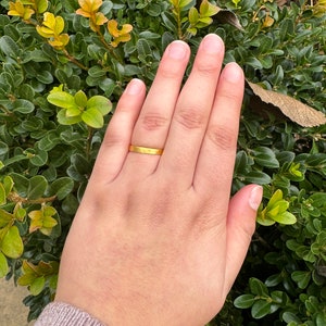Pode incluir: Um close-up de uma m&atilde;o usando uma fina alian&ccedil;a de ouro. A m&atilde;o est&aacute; apoiada em um leito de folhagem verde.