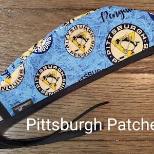 May include: A blue and black surgical scrub hat with a repeating pattern of the Pittsburgh Penguins logo. The hat has a black elastic tie. The text "Pittsburgh Penguins" is printed on the hat.