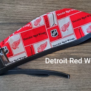May include: Red and white fabric scrub hat with a black trim. The fabric features a repeating pattern of the Detroit Red Wings logo, the NHL logo, and the words "Detroit Red Wings".