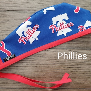 May include: A blue surgical scrub cap with a red trim and ties. The cap features a repeating pattern of the Philadelphia Phillies logo in white and red. The word "Phillies" is printed in white on the cap.