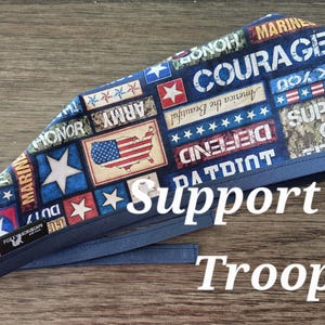 May include: A blue scrub cap with a patriotic design featuring the American flag, stars, and military-themed text. The words "Support Our Troops" are displayed in white. The cap has blue ties.