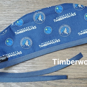 May include: Blue surgical scrub hat with a repeating pattern of the Minnesota Timberwolves logo. The logo features a basketball, a wolf, and the team name.
