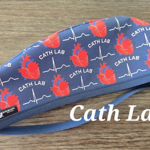 May include: A navy blue surgical cap with a repeating pattern of red anatomical hearts and white EKG lines. The words "CATH LAB" are printed throughout the design. The text "Cath Lab" is printed in white on the front.